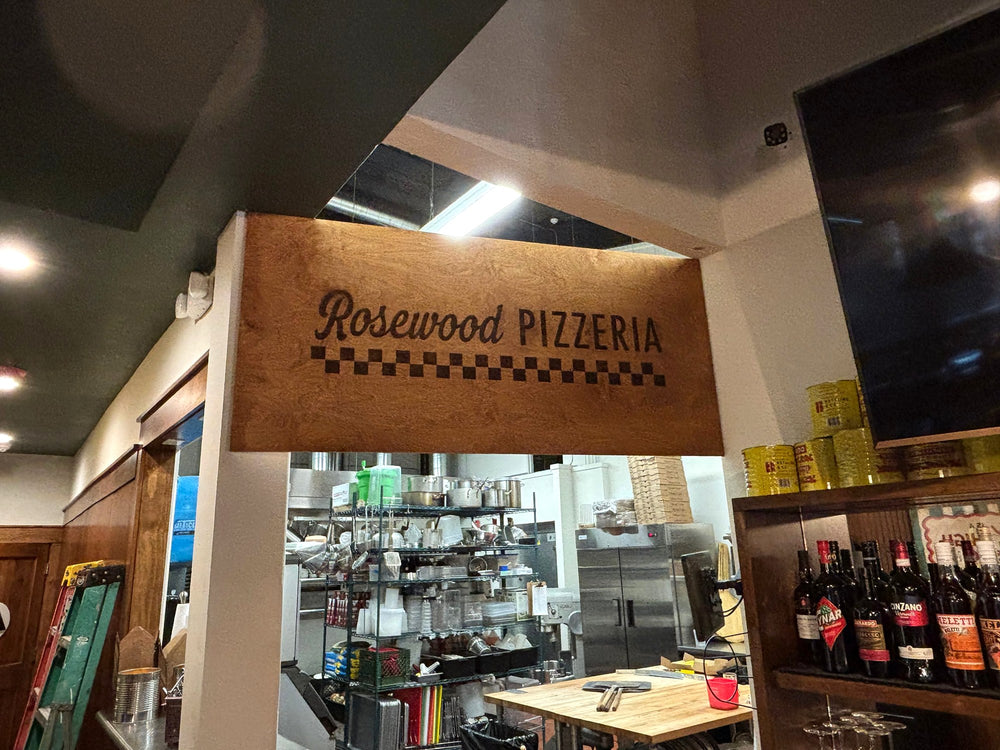 Laser-engraved wood sign reading Rosewood Pizzeria above the kitchen pass-through