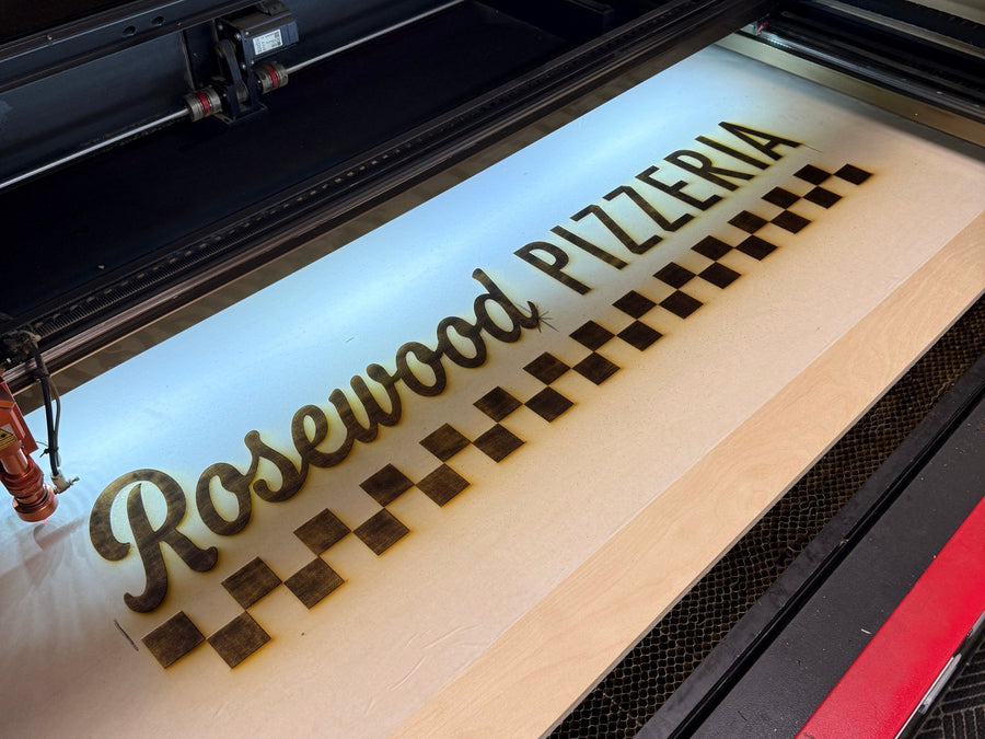 Rosewood Pizzeria lettering and checkerboard being engraved on a wood panel inside a laser engraver
